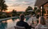 Ein Paar sitzt bei Sonnenuntergang auf einer Terrasse an einem Gartenpool und trinkt Wein.