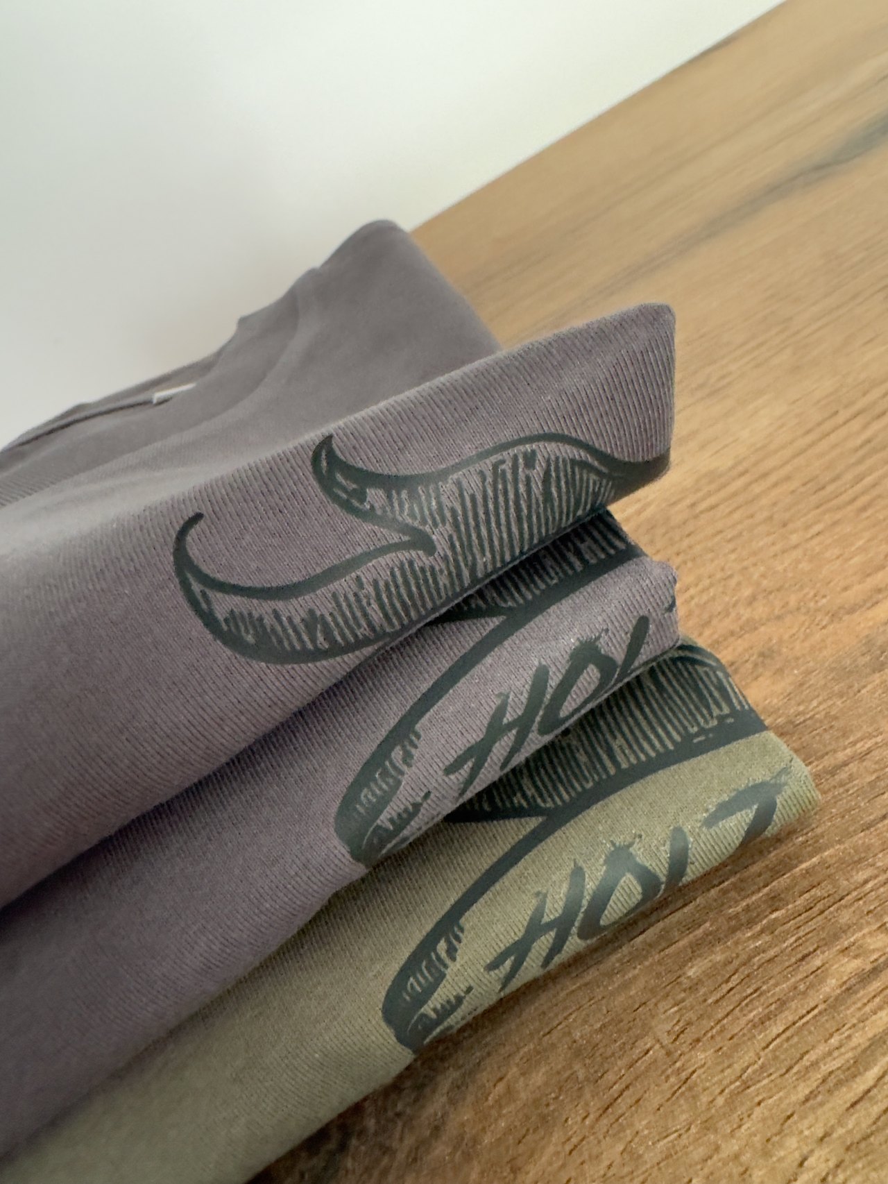 Stapel grauer und grüner T-Shirts mit schwarzem HOLZ-Schriftzug