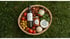 A woven basket filled with olive oil, tomatoes, basil, nuts, pesto, a jar of sauce, and cheese on grass.