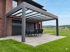 Moderne Terrassenüberdachung aus grauem Metall mit Glasdach, darunter ein Esstisch und Stühle auf einer gepflasterten Terrasse.