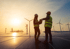 Zwei Ingenieure stehen auf Solarmodulen vor Windkraftanlagen bei Sonnenuntergang.