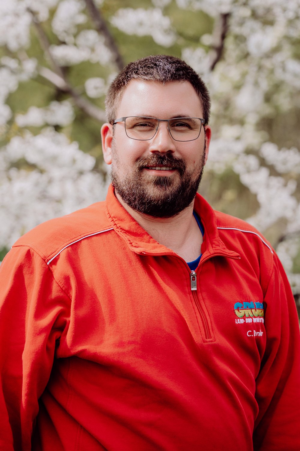 Mann mit Bart und Brille in rotem Pullover mit Reißverschluss und "GREEN LAND-PRO MOWING"-Logo vor weißen Blumen.