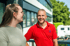 Ein lachender Mann in einem roten Poloshirt unterhält sich im Freien mit einer Frau vor einem Bürogebäude.