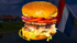 Großer Cheeseburger mit zwei Patties vor einer niederländischen Flagge und Häusern in einer Küstenlandschaft.