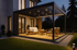 Moderne Pergola mit Lamellendach und Lichtern über einer Holzterrasse mit Gartenmöbeln bei Dämmerung.