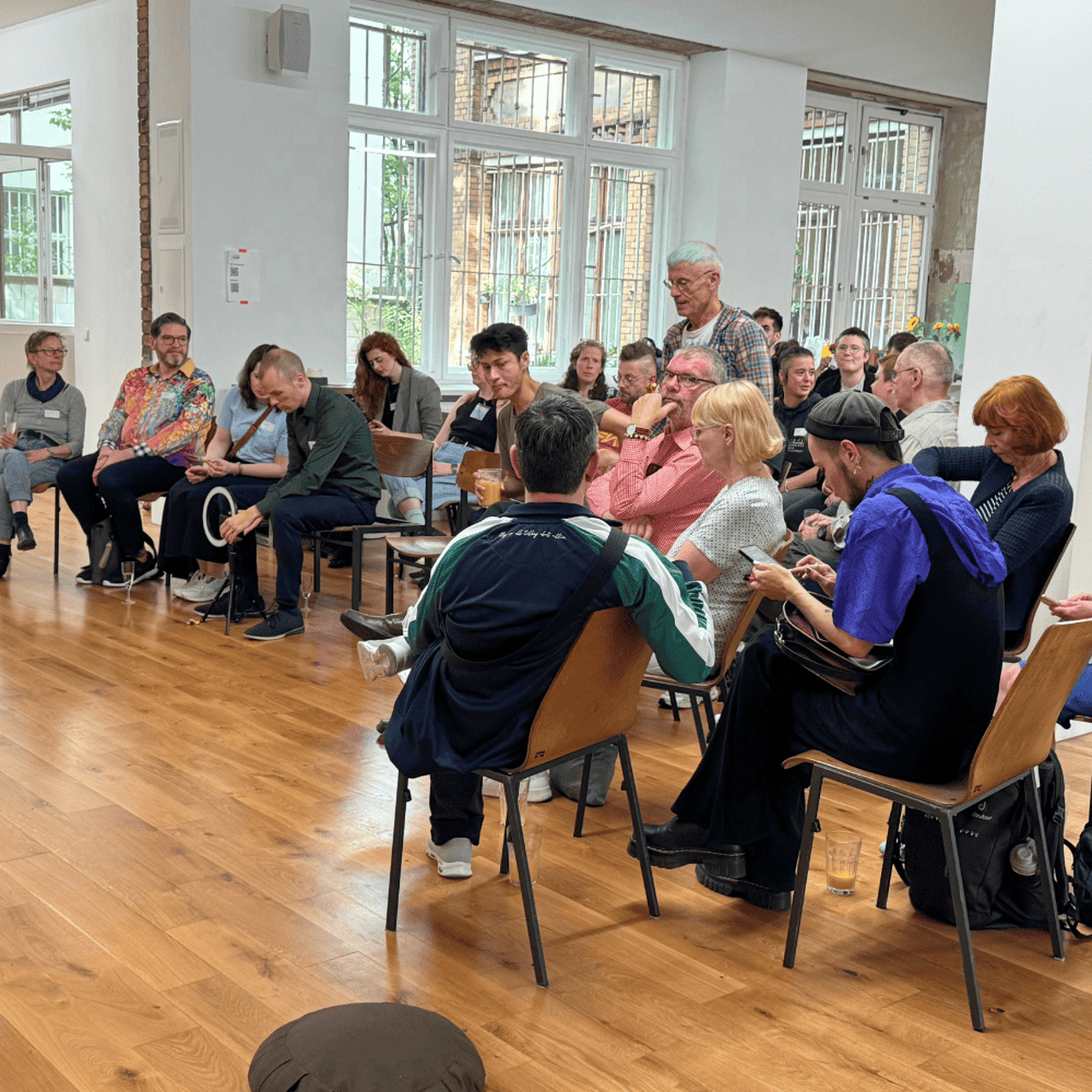 A group of people sit on chairs in a large room with a wooden floor and windows.