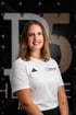 Lächelnde Frau mit braunen Haaren im weißen Sportshirt mit dem Logo THERAPIER und Pier5 vor dunklem Hintergrund.