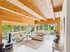 Modernes Wohnzimmer in einem Holzhaus mit massiven Deckenbalken, hellen Sofas, Couchtisch und großen Fensterfronten zum Garten.