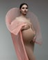 Schwangere Frau in engem, beigem Kleid mit einem großen, fächerartigen rosa Plissee-Stoffelement vor grauem Hintergrund.