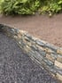 Natursteinmauer aus grauen und braunen Steinen trennt eine Schotterfläche von einem bepflanzten Hang.