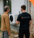 Ein Techniker mit "Das ElektroWerk"-Shirt erklärt einem Mann eine Wärmepumpe an einer Hauswand.