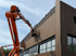 Orangefarbener Gelenksteiger neben einem Gebäude mit Holzverkleidung und Fenstern unter blauem Himmel.