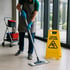 Reinigungskraft wischt Boden mit Mopp; gelbes "CAUTION WET FLOOR"-Schild steht daneben.