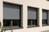 Drei Fenster mit geschlossenen grauen Rollläden an einer hellen Hauswand.
