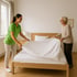 Haushalt, Lachende Pflegerin und Seniorin machen gemeinsam ein Bett mit weißer Bettwäsche. 