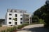 Modernes, weißes Mehrfamilienhaus mit grauen Dachgeschossen und Balkonen, umgeben von Bäumen und einer Straße.