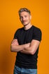 Ein lächelnder Mann mit blonden Haaren, schwarzem T-Shirt und blauen Jeans steht mit verschränkten Armen vor orangefarbenem Hintergrund.