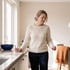 A woman in a cream sweater stands in a kitchen holding a glass of water on a countertop.