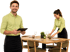 Kellner mit Tablet lächelt, während eine Kollegin im Hintergrund einen Restauranttisch deckt.