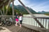 Zwei Frauen mit Fahrrädern stehen auf einer Holzbrücke und blicken auf einen Fluss und Berge.