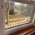 Blick aus einem Fenster auf einen Garten mit Bäumen, einer Hecke und einem roten Backsteingebäude im Hintergrund.