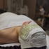 Eine Frau liegt entspannt mit einer grünen Jade-Augenmaske und einem weißen Handtuch um den Kopf.