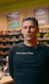 Ein Mann in einem dunkelblauen Hemd spricht in die Kamera; im Hintergrund sind Sportschuhe und die Aufschrift "Firma Sport Flöss" zu sehen.