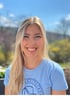 Lächelnde blonde Frau in hellblauem T-Shirt mit „GESUNDHEITSPARK“-Logo, im Freien.