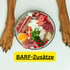 Hundepfoten um eine Schüssel mit rohem Fleisch, Ei, Beeren und Gemüse; Text: BARF-Zusätze.