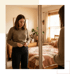 A woman in a brown sweater and dark pants looks in a mirror while struggling to fasten her waistband in a bedroom.