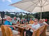Eine Gruppe älterer Menschen sitzt an einem Holztisch unter einem Veltins-Sonnenschirm im Freien und trinkt Bier.