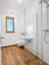 Modernes Badezimmer mit Holzboden, Glasdusche, weißem Waschbecken und wandhängendem WC unter einem Holzfenster.