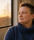 Porträt einer Person mit kurzem Haar und blauem Rollkragenpullover, die aus einem Fenster in das Abendlicht blickt.