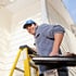 Lachender Handwerker mit blauer Kappe steht auf einer gelben Leiter an einer hellen Hausfassade und hält eine Werkzeugschale.