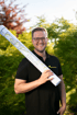 Lächelnder Mann mit Brille hält eine Planrolle von Garten Schmid vor grünem Hintergrund.