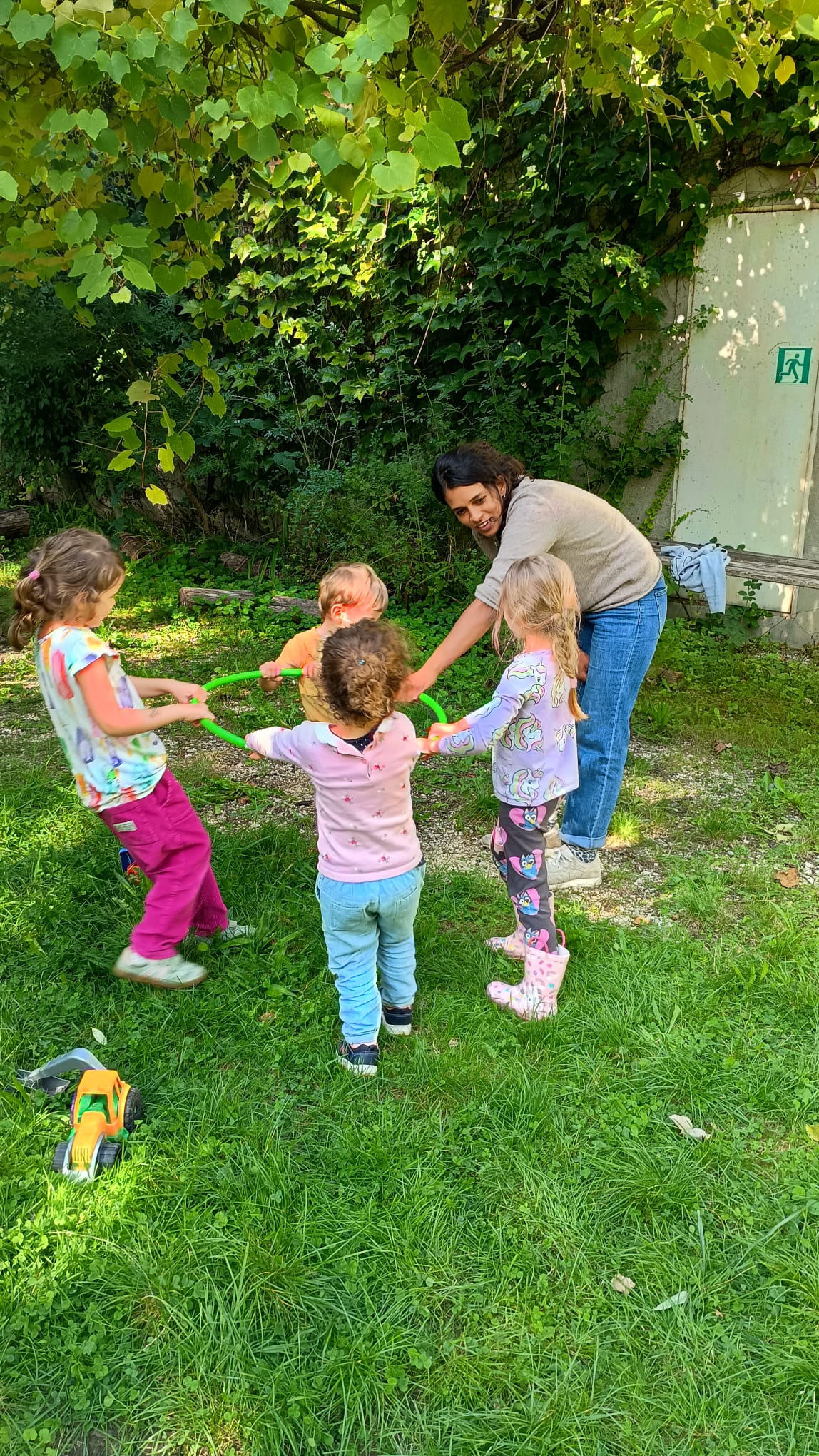 Betreuungsperson und Kinder spielen gemeinsam mit einem grünen Reifen im Garten.