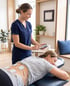 A physical therapist in blue scrubs uses a handheld TENS unit on a patient lying prone with electrodes on her lower back.