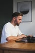 Mann mit Bart, weißem T-Shirt, tippt auf Laptop an einem Holztisch, im Hintergrund ein gerahmtes Bild.