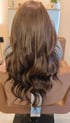 Rückansicht einer Person mit langen, glänzenden braunen Haaren mit leichten Wellen, die auf einem Friseurstuhl sitzt.