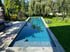 Schmaler, rechteckiger Pool mit blauem Wasser in einem Garten mit Rasen, Bäumen und einer Holzterrasse.