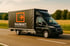 Schwarzer Raumakt-LKW mit orangefarbenem Logo fährt auf einer Landstraße.