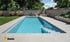 Ein rechteckiger Pool mit klar blauem Wasser, umgeben von grauen Fliesen, Garten und Steinmauer im Hintergrund.