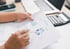 Hände halten Finanzbericht mit Diagrammen und zeigen mit einem Stift auf ein Kreisdiagramm; Laptop und Taschenrechner im Hintergrund.