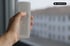 Hand hält eine weiße Fernbedienung mit Pfeiltasten vor einem Fenster mit unscharfem Stadtbild.