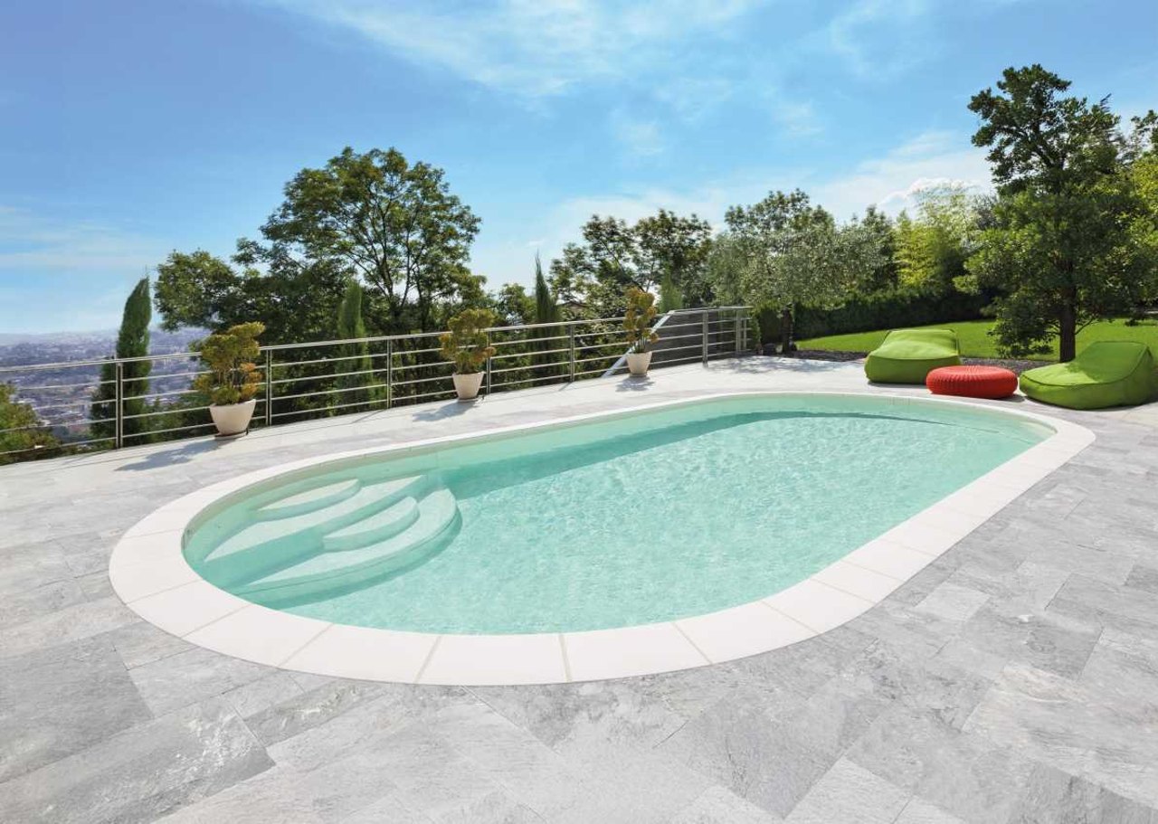 Ovaler Pool mit Treppe, umgeben von grauen Fliesen und Bäumen unter blauem Himmel.