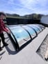 Ein Mann schiebt eine flache, transparente Poolüberdachung mit schwarzem Rahmen über einen Gartenpool.