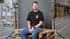 Mann in schwarzem T-Shirt mit "Order Picker"-Logo sitzt auf Holzpalette vor grauem Hintergrund in Lagerhalle.