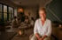 Lächelnde Frau im Vordergrund eines Meditationsraums mit einer im Hintergrund sitzenden Frauengruppe bei Kerzenschein.