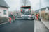 Straßenfertiger und Bauarbeiter in orangefarbener Arbeitskleidung asphaltieren eine neue Straße vor Häusern.