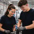 Zwei junge Arbeiter in Schutzbrillen und Handschuhen montieren ein Metallteil in einer Fabrikhalle.